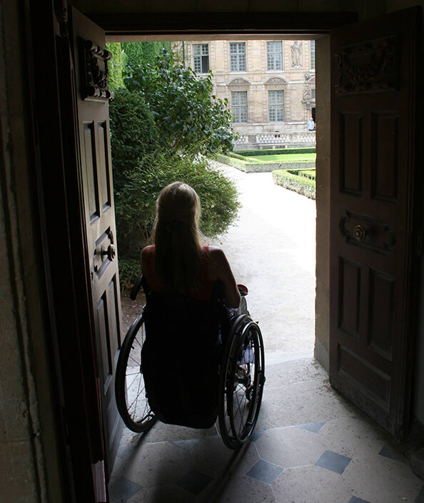 Woman in wheelchair sits in dark room looking out door to outside courtyard