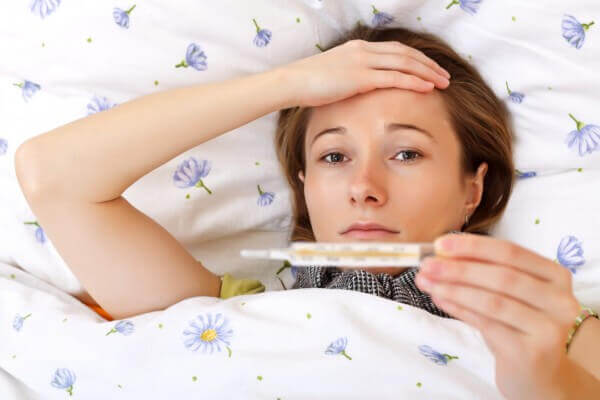 image of a girl laying on a bed with one hand on her forehead while holding a thermometer from other hand