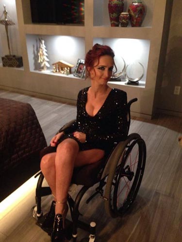 A woman in a wheelchair, wearing a black dress with her legs crossed over.