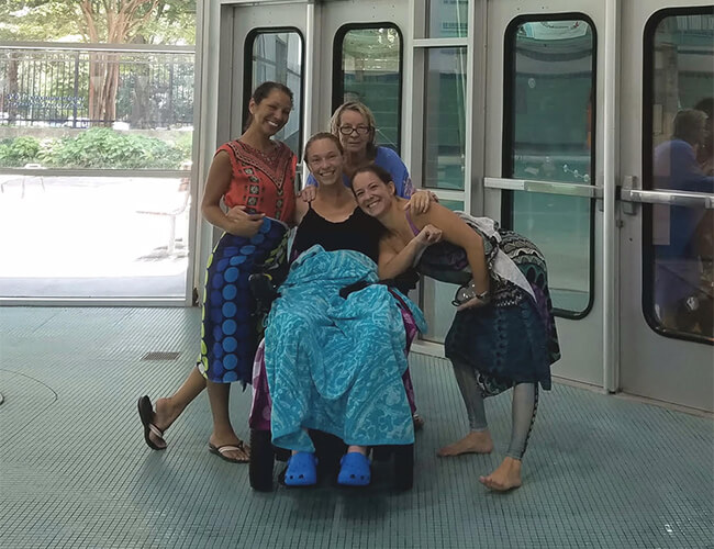 three women sitting at swim area standing and smiling with a disabled women sitting on a wheelchair