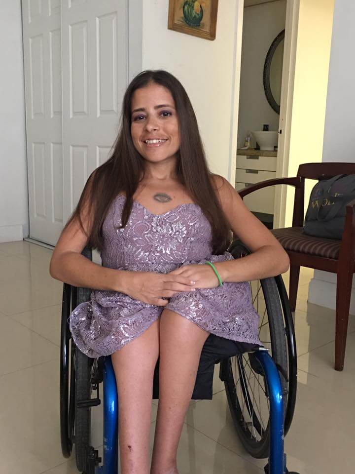 Kat sitting in her wheelchair in a sparkly violet short dress smiling at the camera