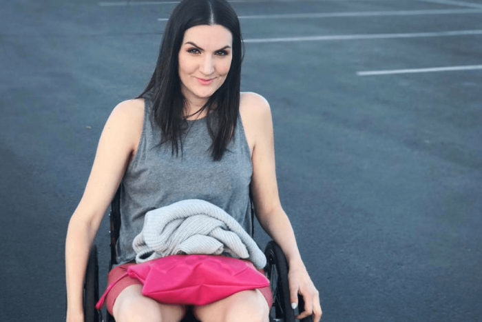 Gina Schuh in her wheelchair. looking at camera in a parking lot with pink shorts and grey tank top